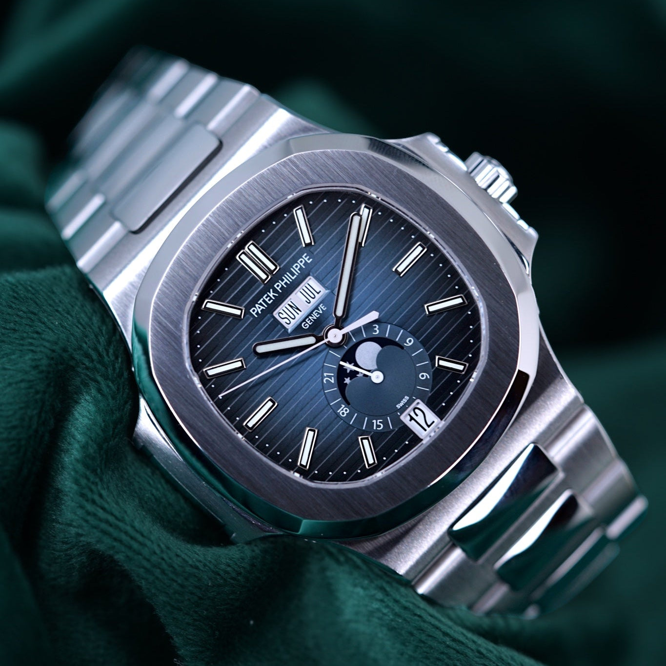 A stainless steel wristwatch with a dark blue sunburst dial and a matching metal bracelet, resting on draped dark green fabric.